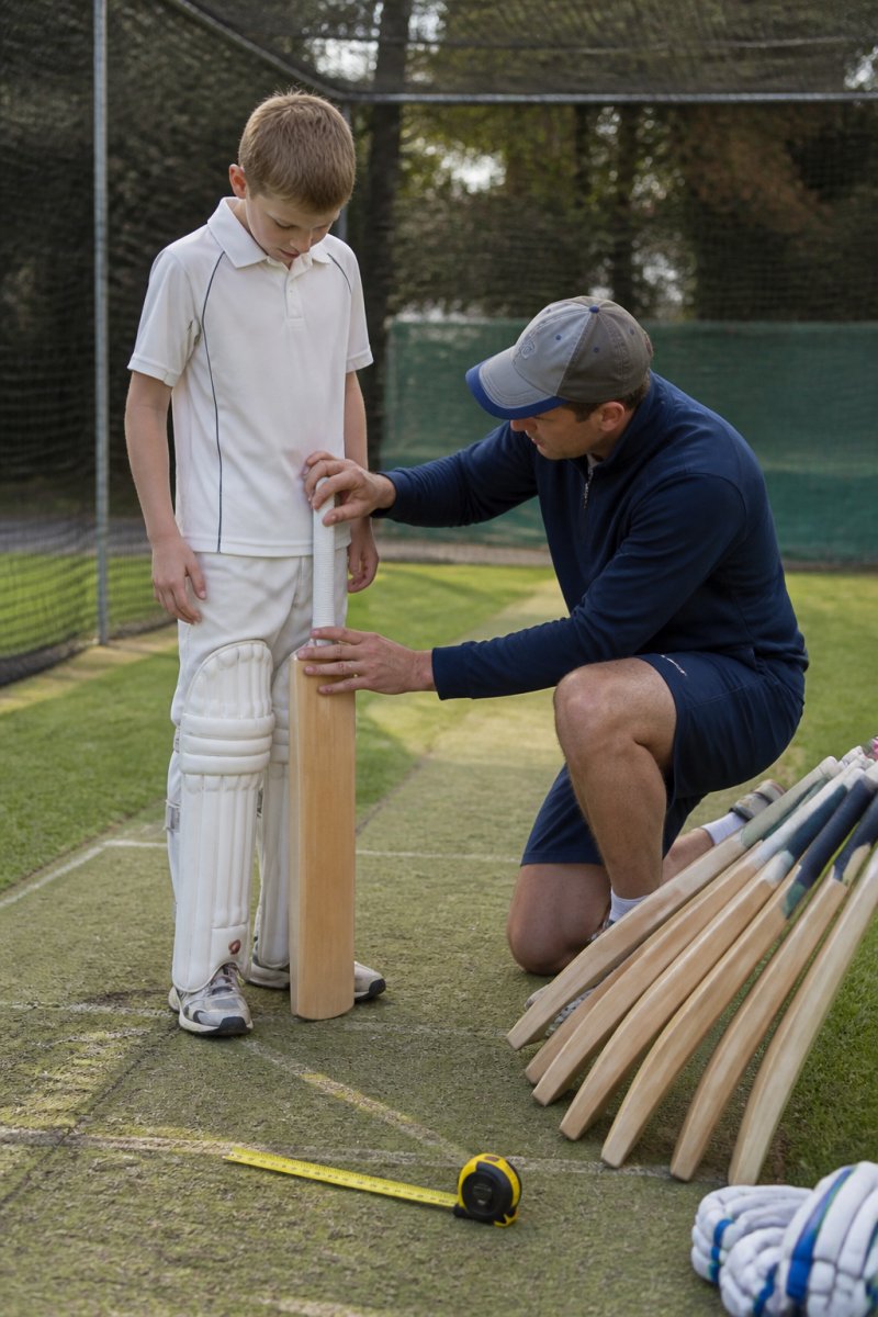 A cricket coach kneeling beside a young player on a practice pitch, holding a full-size bat against the player's leg to check the height, with a measuring tape and a row of bats of different sizes resting on the grass nearby