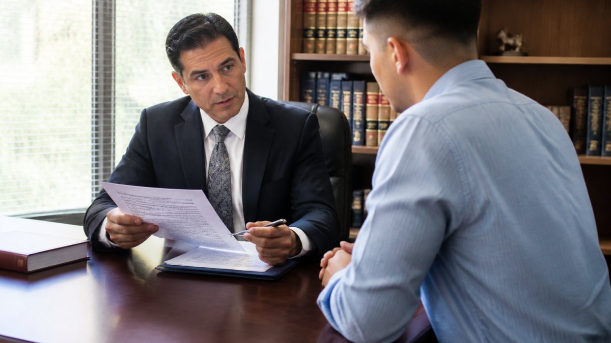 Criminal defense attorney in professional office setting meeting with client across desk, reviewing case documents and discussing legal strategy with natural office lighting