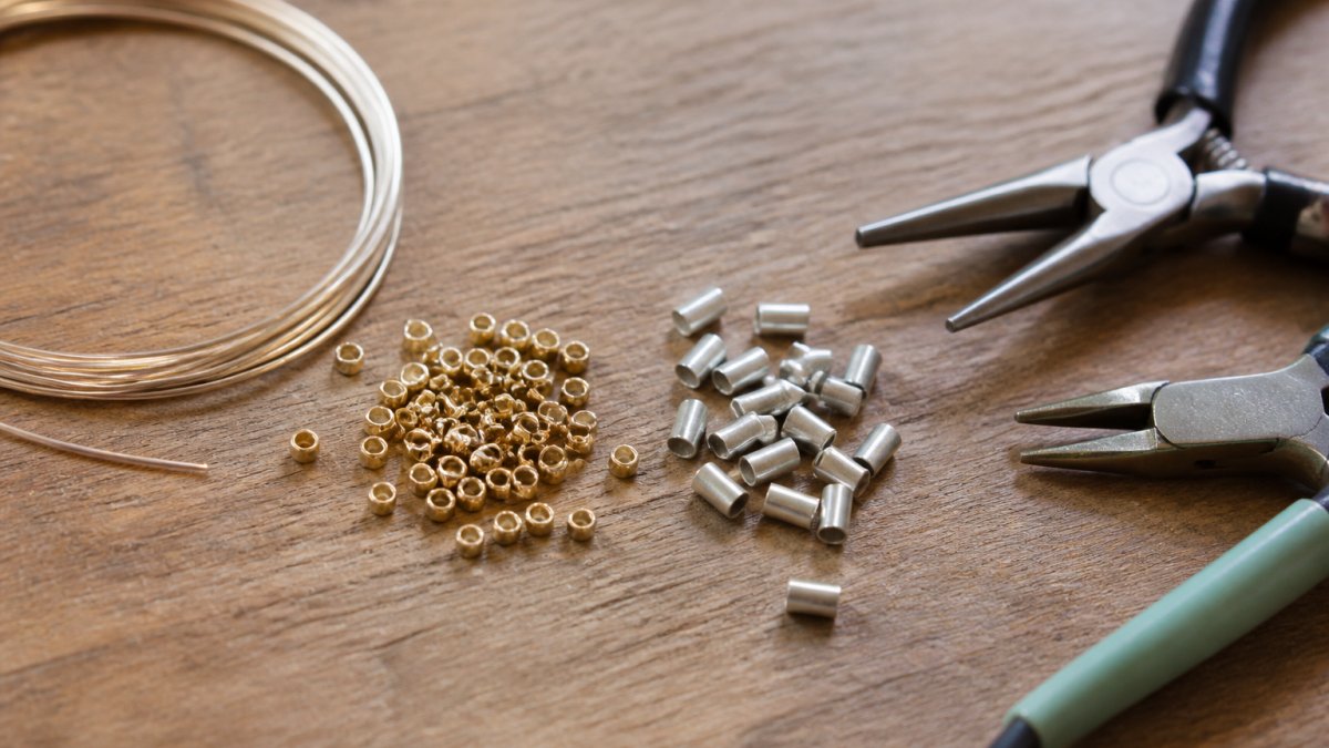 Close-up on a craft table showing a small pile of round crimp beads next to a row of cylindrical crimp tubes, with fine beading wire and jewelry pliers resting beside them under warm daylight
