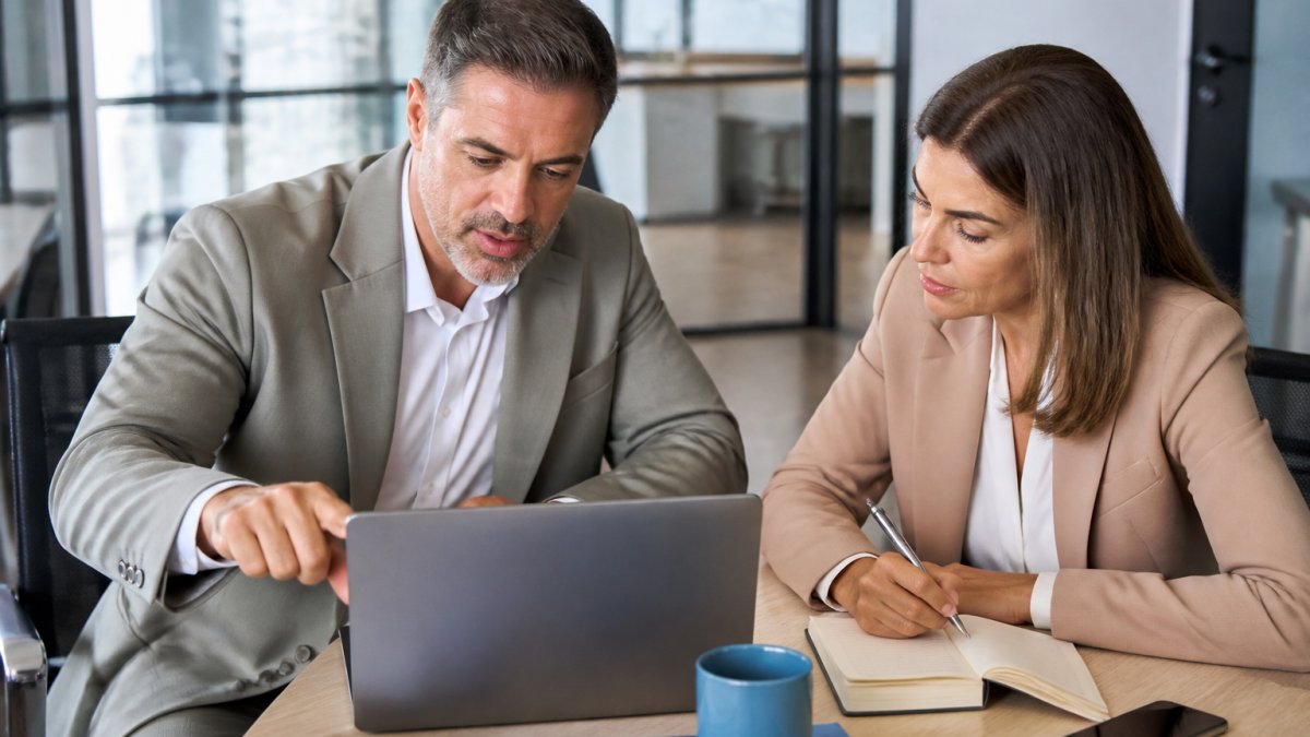 Two business professionals in a bright office reviewing a CRM dashboard on a laptop, one pointing at the screen while the other takes notes, suggesting a strategy discussion