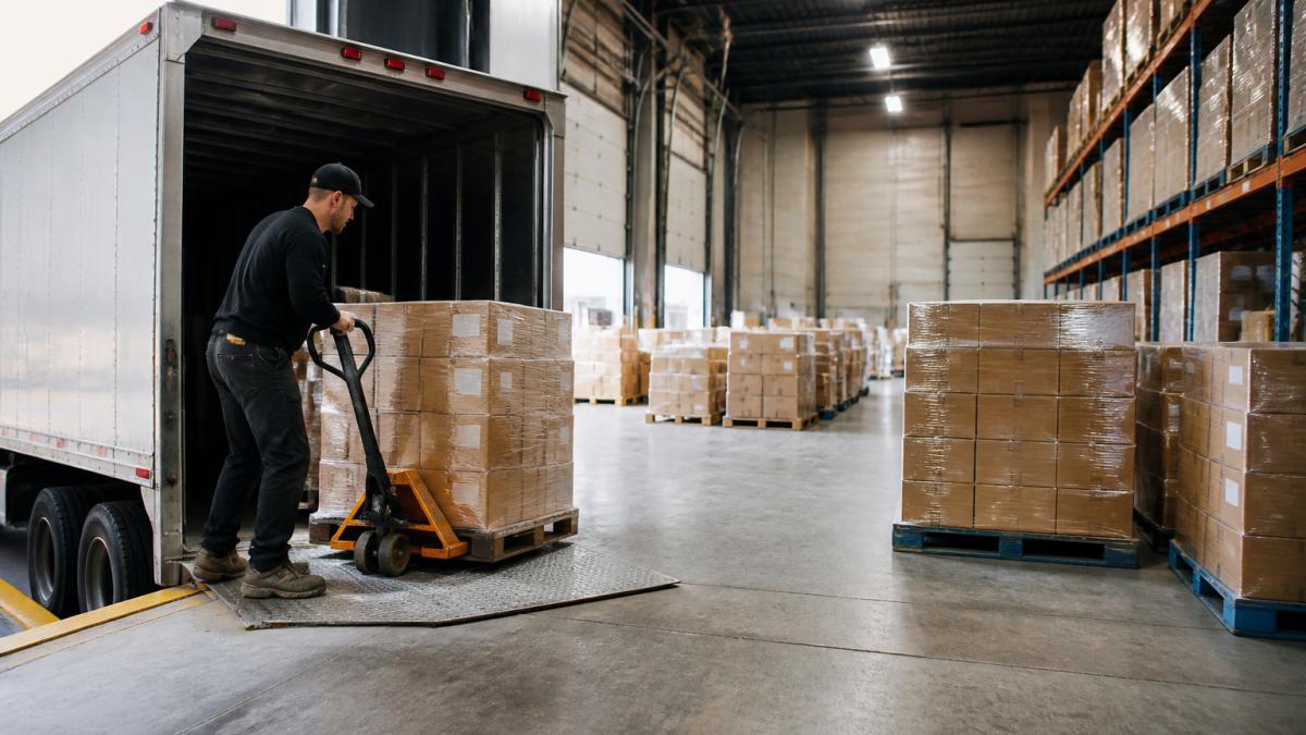Warehouse workers unloading pallets from a truck at a dock door, with organized freight staging areas and sorted shipments visible in the background under bright overhead lighting