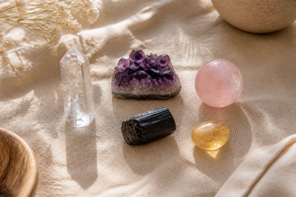 A flat-lay arrangement of five beginner crystals, clear quartz point, amethyst cluster, rose quartz sphere, black tourmaline rough, and citrine tumble, arranged on a natural linen cloth with soft afternoon window light casting gentle shadows
