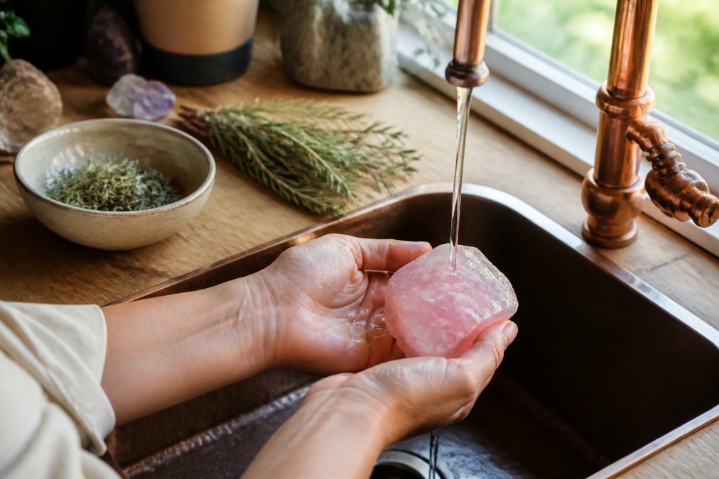 A pair of hands gently holding a rose quartz crystal under a thin stream of water from a copper faucet, with dried rosemary and a small ceramic bowl visible on the wooden surface beside the sink, soft natural daylight coming through a nearby window