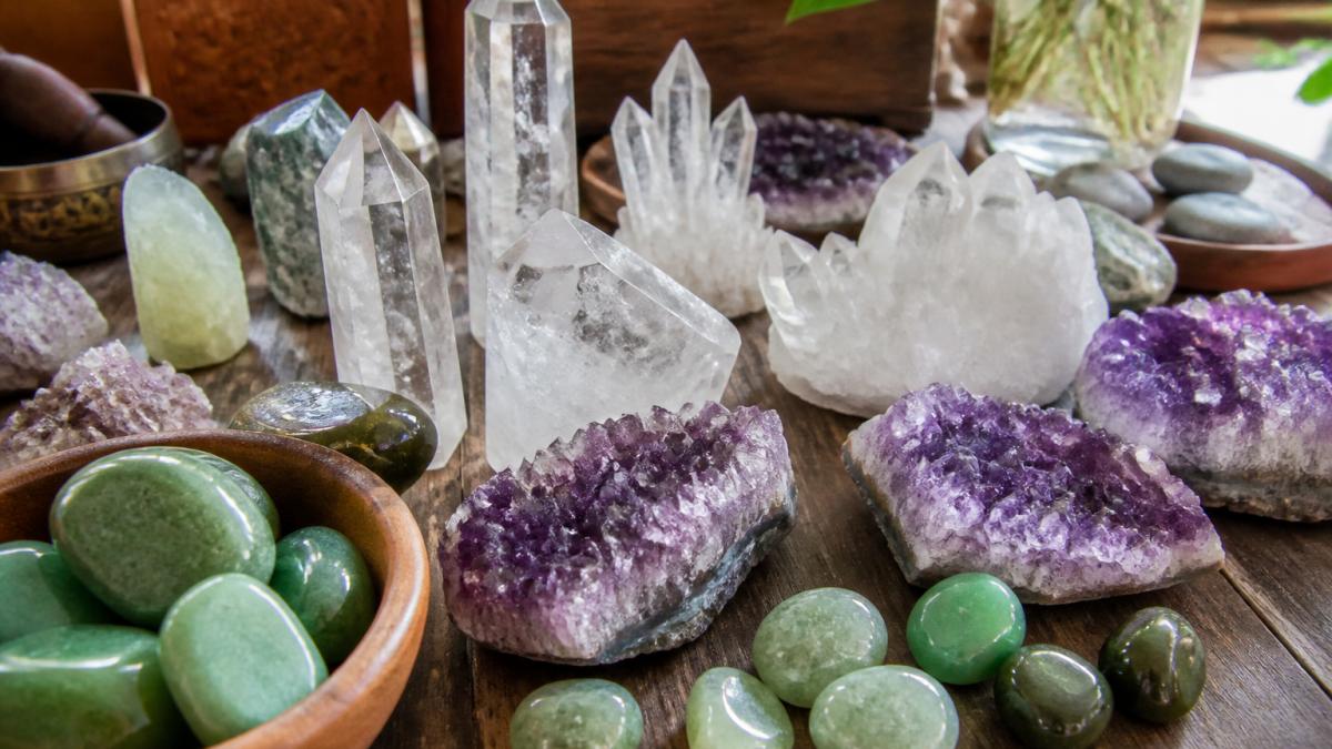 Close-up arrangement of various raw and polished crystals including clear quartz points, jade tumbles, green aventurine, and amethyst clusters displayed on natural wood surface with soft natural lighting highlighting their distinct colors and textures