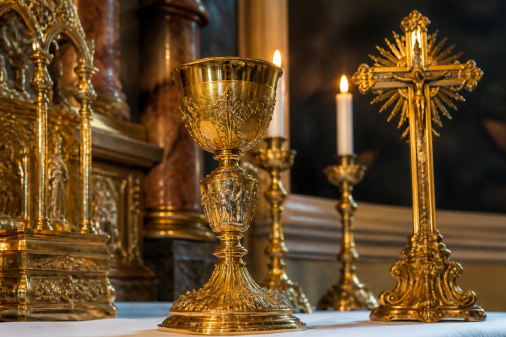 Close-up of ornate golden religious artifacts including a golden chalice, ornamental cross, and temple decorations illuminated by soft candlelight in a sacred space