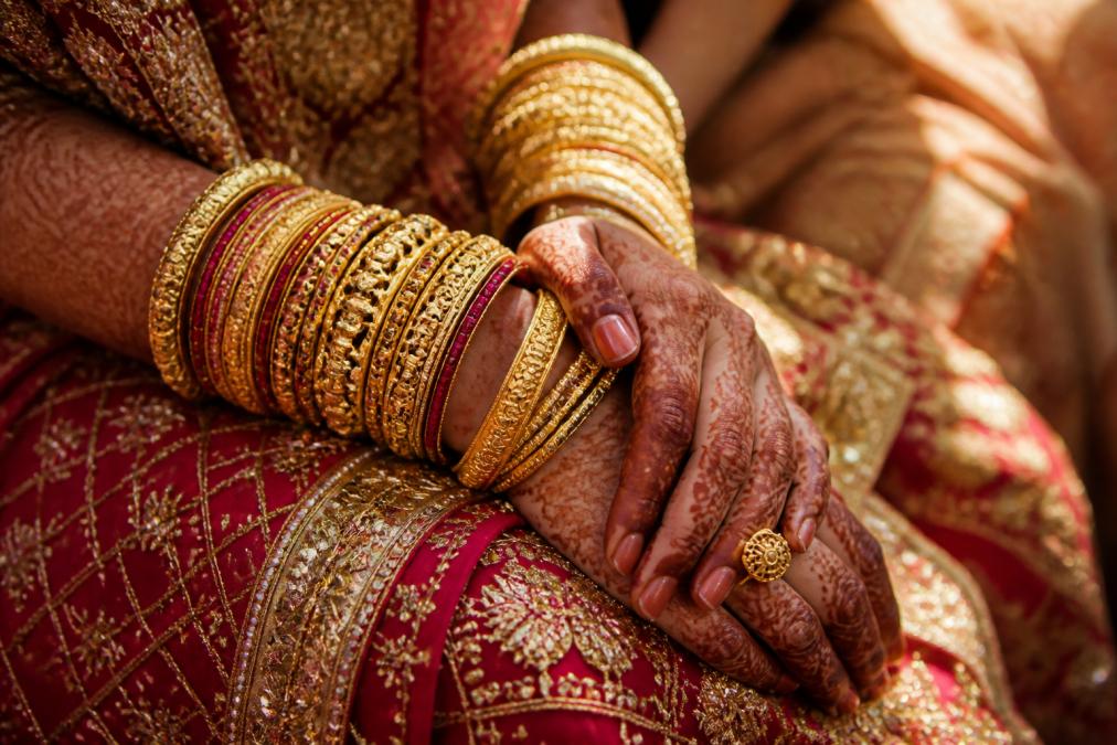Hands of a bride adorned with intricate traditional gold jewelry and bangles during a wedding ceremony, showing detailed craftsmanship with natural sunlight illuminating the metal