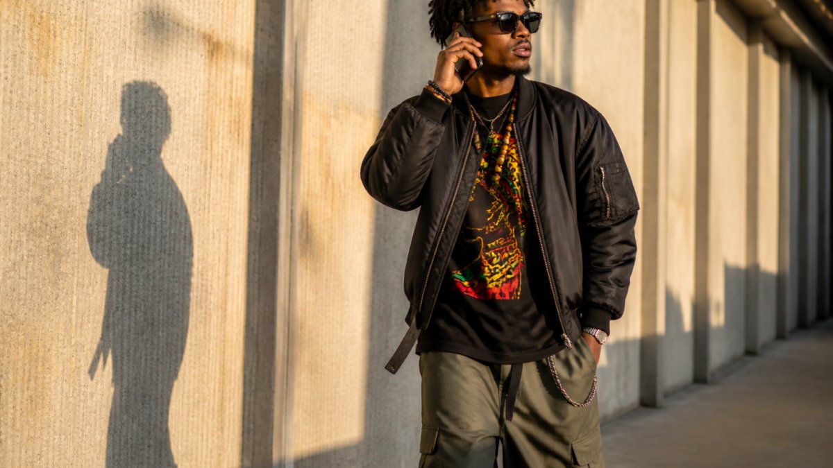 Young adult wearing layered cultural streetwear with bold graphic tee, structured bomber jacket, and statement accessories, posing confidently in urban setting with natural morning light casting shadows