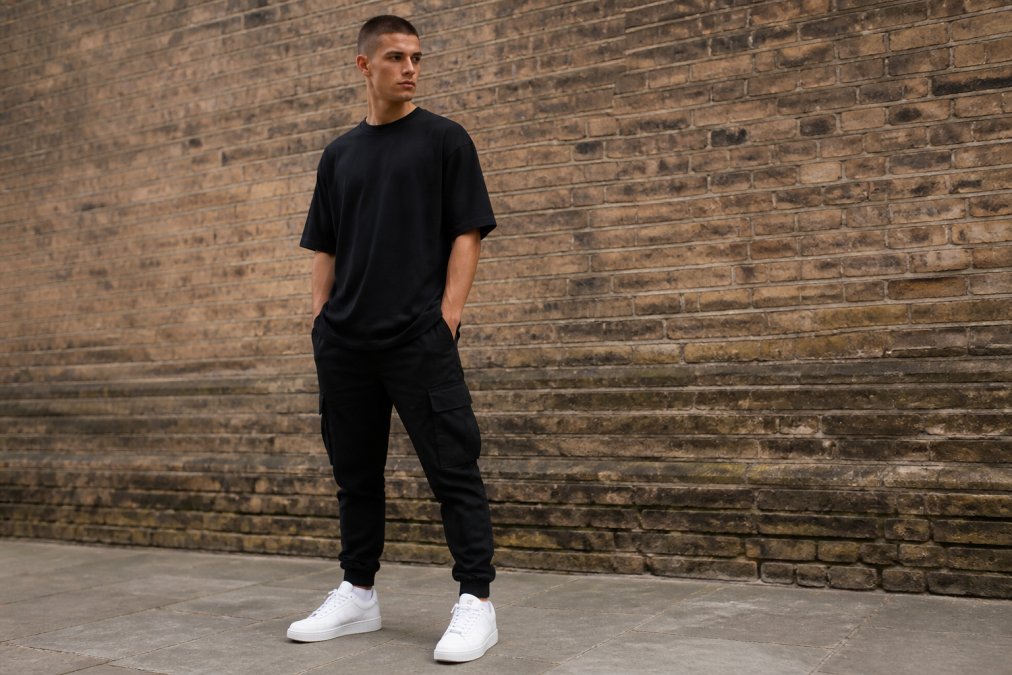 Young man wearing an oversized black tee styled with tapered black cargo pants and white low-top trainers, photographed from full-body angle against a neutral urban brick backdrop with natural daylight