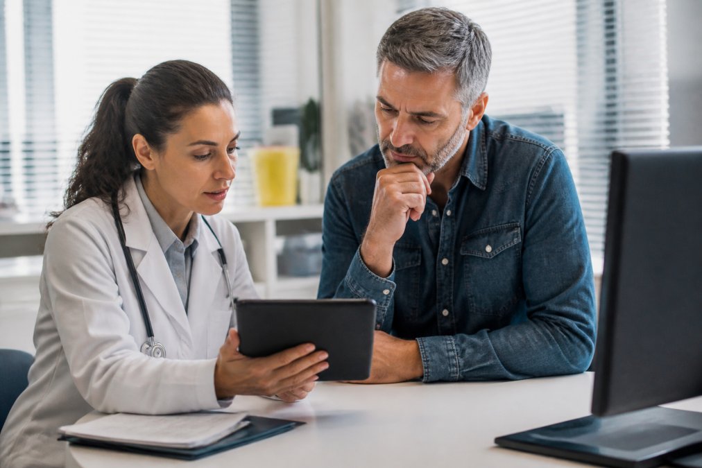 A female physician in a white coat reviewing a digital chart with a middle-aged male patient in a bright, modern consultation room