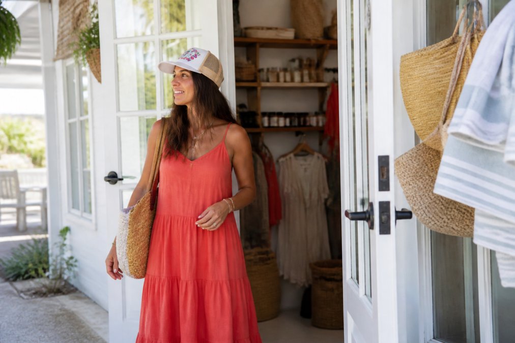A woman wearing a flowing coral maxi dress and a canvas embroidered trucker hat, standing near a bright coastal boutique entrance with warm natural light and a relaxed beach-town atmosphere