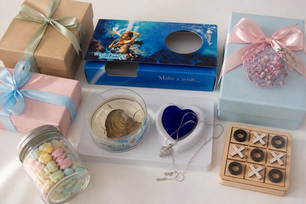 Flat lay of several beautifully wrapped gift boxes and small gift items arranged on a white surface, including a pearl jewelry box, a small game, and a glass jar with folded notes inside