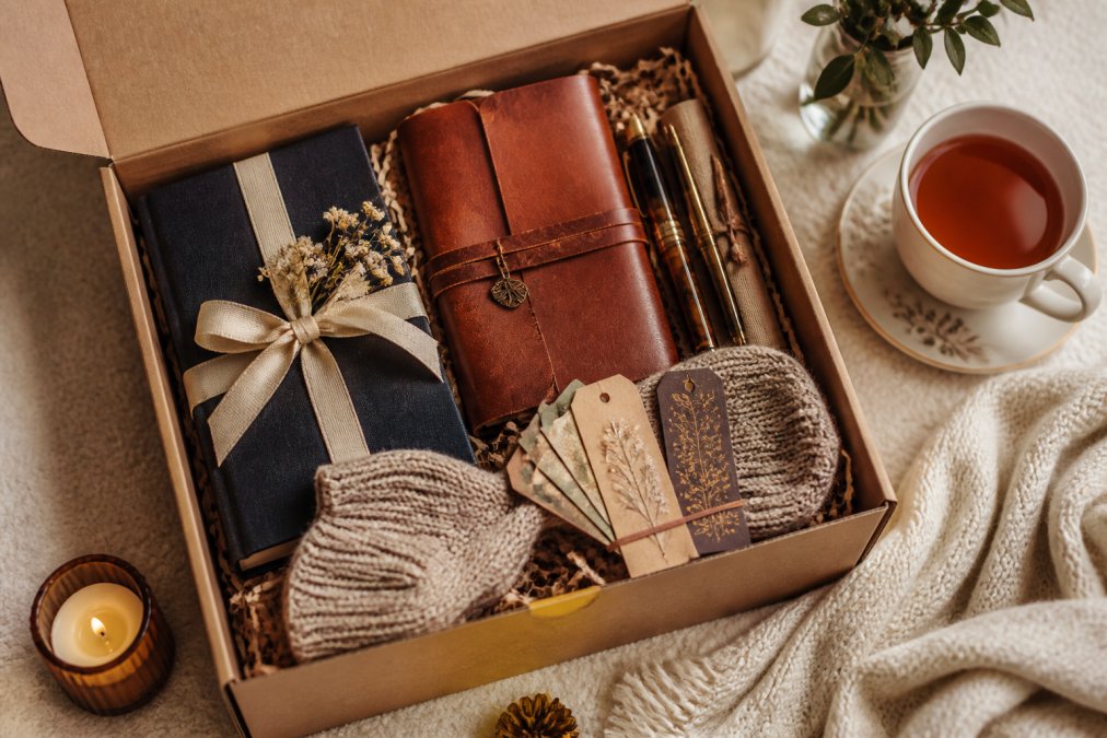 Overhead shot of an open gift box containing a wrapped hardcover book, leather journal, quality pens, bookmarks, and cozy reading accessories arranged artfully on a soft cream surface with warm golden lighting and a cup of tea beside it