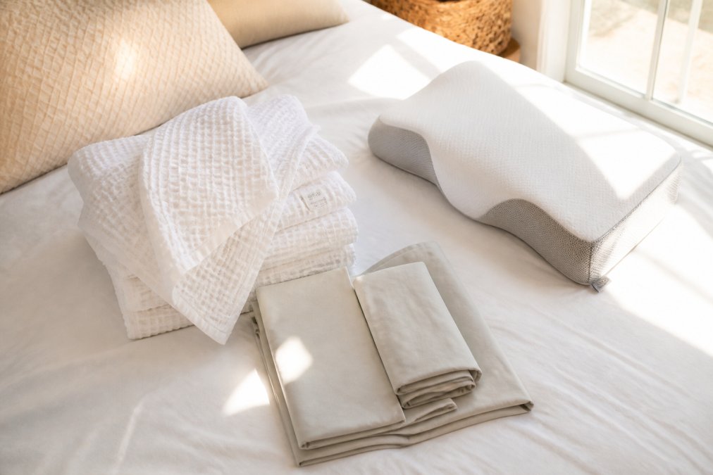 Overhead flat lay of soft folded towels in neutral tones, cooling bamboo sheets, and an adjustable pillow arranged on a made bed with warm natural light streaming through a bedroom window