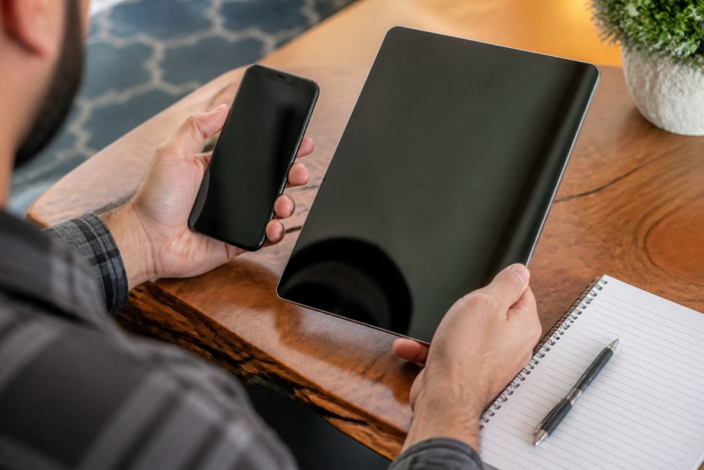 Close-up of hands holding a smartphone and tablet side-by-side, displaying product specifications and reviews on the screens, with a notebook and pen nearby for note-taking