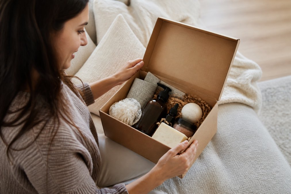 Person opening a wellness delivery box at home, showing organized wellness products and care items inside with natural lighting