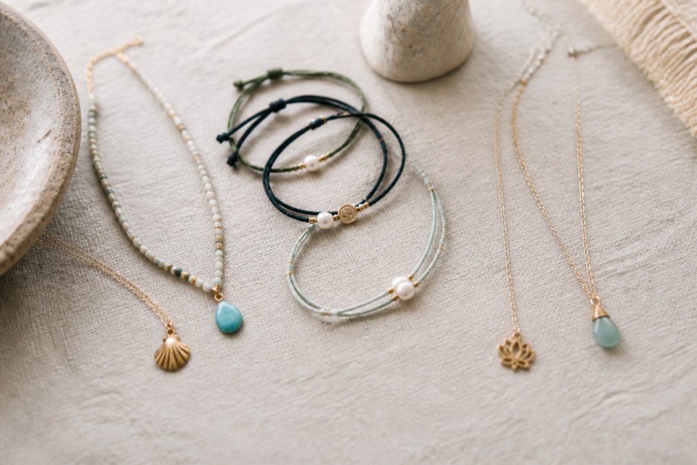 Flat lay of handcrafted necklaces and bracelets on a light linen surface with soft natural light, showing delicate charms and cord details