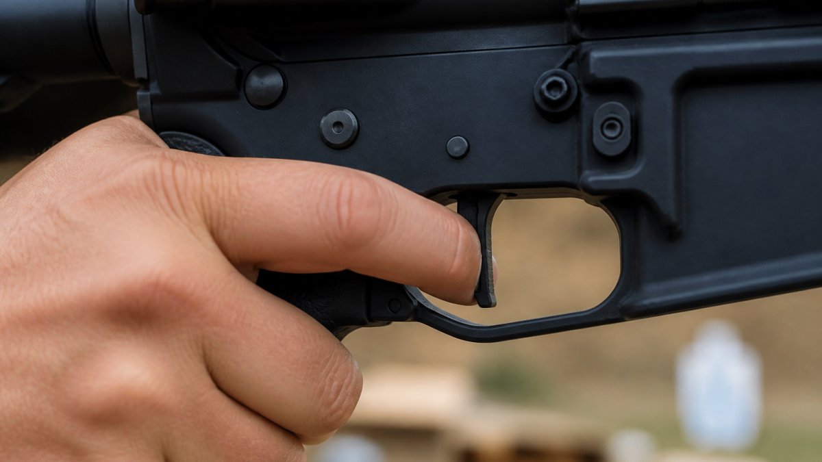 Close-up of a shooter's index finger resting on a flat-faced trigger inside an AR-15 trigger guard, range environment blurred in background