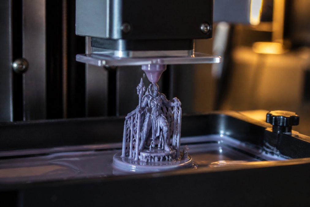Close-up of a 3D printer nozzle laying down fine resin layers to create a detailed miniature figurine, with soft workshop lighting in the background