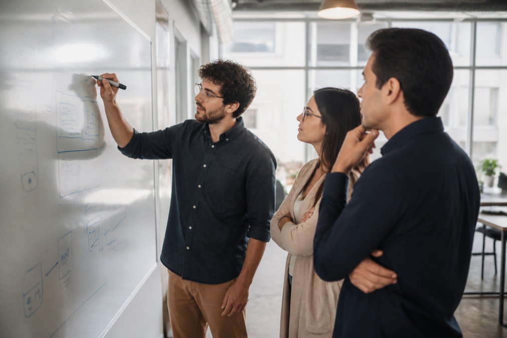Professional software developers and product managers collaborating at a whiteboard, sketching chatbot architecture and conversation flows during a planning session in a modern office with natural lighting