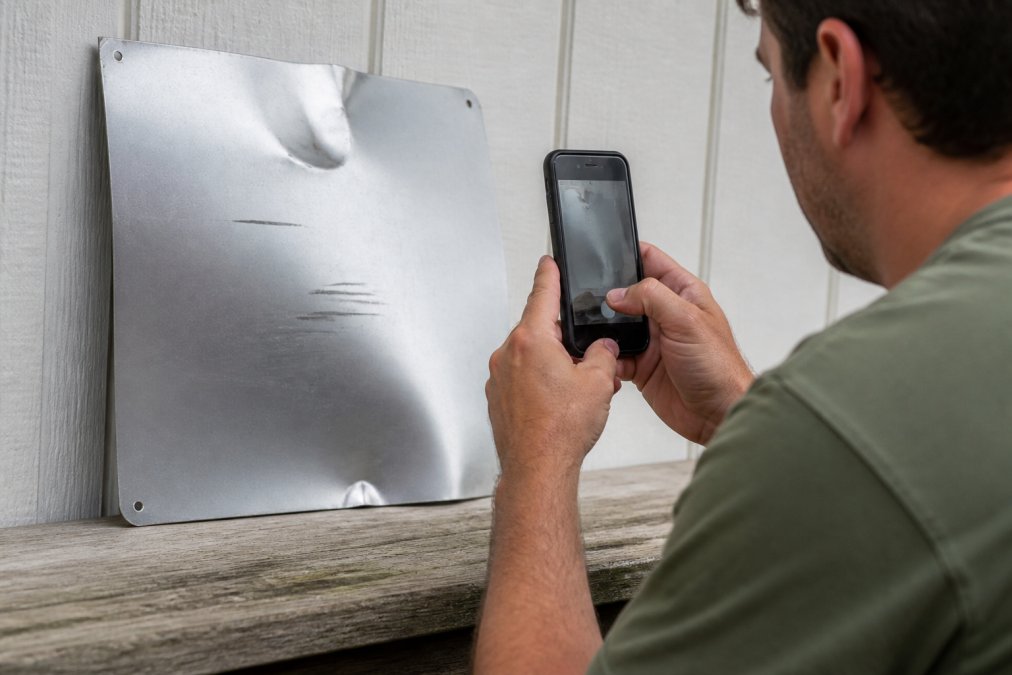 Homeowner photographing a damaged custom aluminum sign with a smartphone, capturing the dent and scratches from multiple angles against a white wall in natural daylight