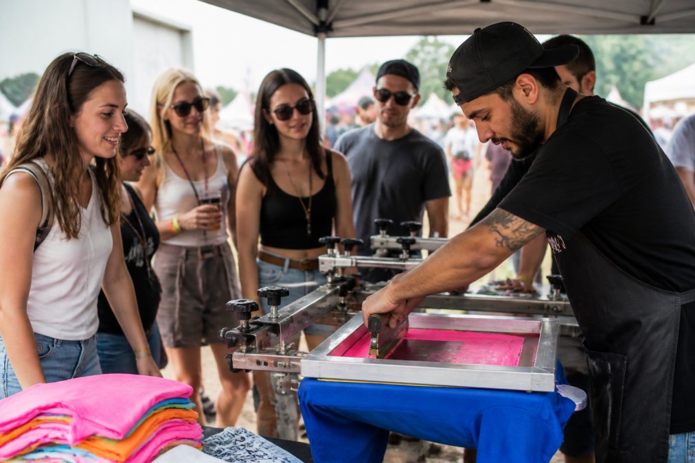 Professional screen printing operator setting up and operating custom apparel printing equipment at outdoor event venue, with attendees watching colorful shirts being printed in real-time under bright natural lighting