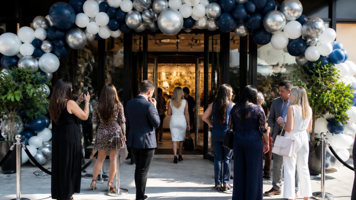 Wide-angle view of an elegant storefront entryway decorated with a custom balloon arch in sophisticated brand colors, with well-dressed guests admiring the display and taking photos