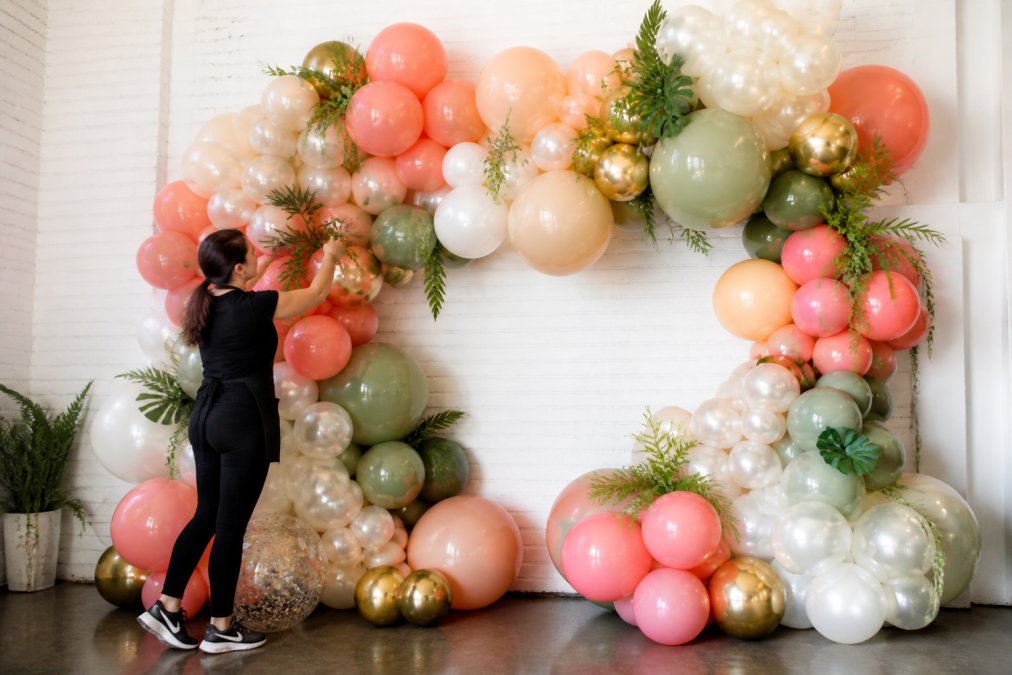 A professional balloon artist arranging a colorful organic balloon garland backdrop against a white wall, with foil balloons and latex balloons of varying sizes creating a layered, handcrafted display in a bright event space