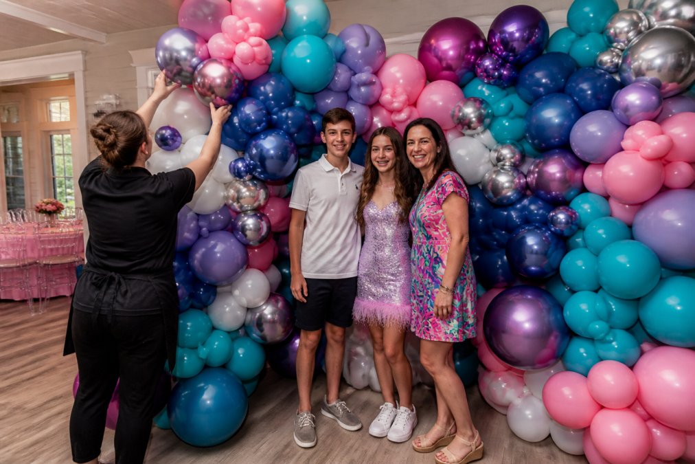 Professional balloon artist arranging colorful custom balloon mosaic backdrop with organic clusters and shimmer accents at a birthday celebration, with guests posing in front for photos
