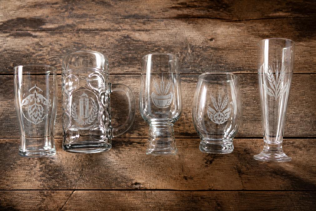 Flat-lay arrangement of five custom-branded beer glasses in different styles, pint glass, stein, schooner, pot, and pilsner glass, each engraved with a logo, arranged on a worn rustic timber surface under warm overhead lighting