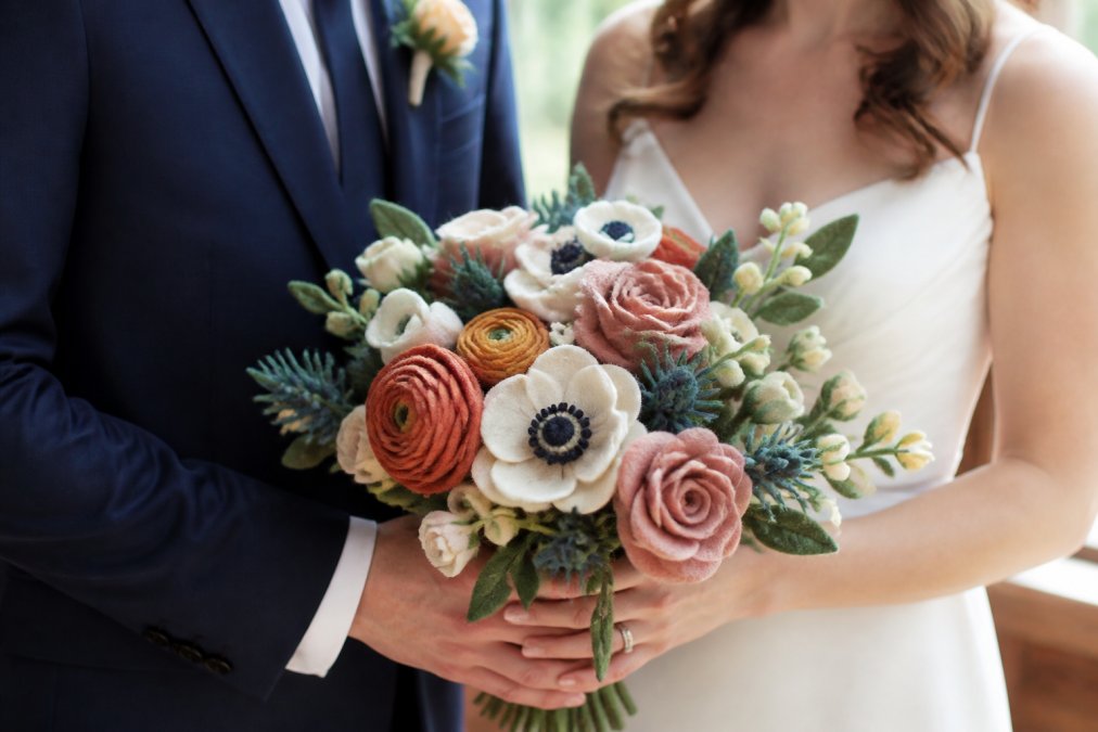 Close-up of a bride and groom holding a handcrafted felt bouquet at a wedding, with soft natural light and a warm, romantic atmosphere