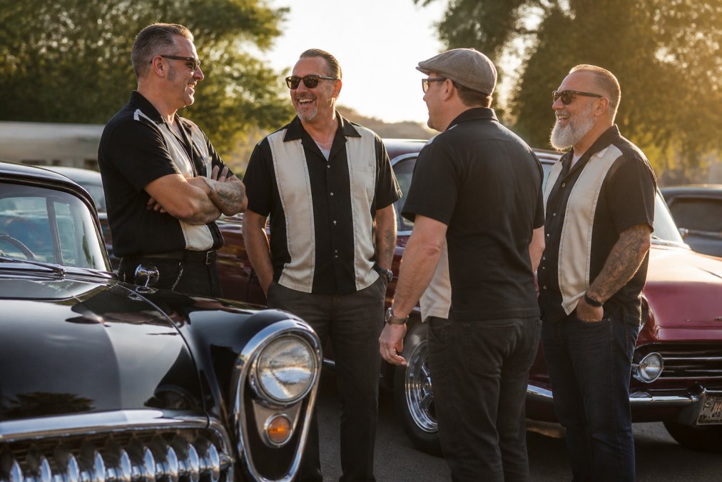 A group of car club members in matching vintage-style bowling shirts standing beside polished classic cars at an outdoor meet, laughing and talking in late afternoon sunlight