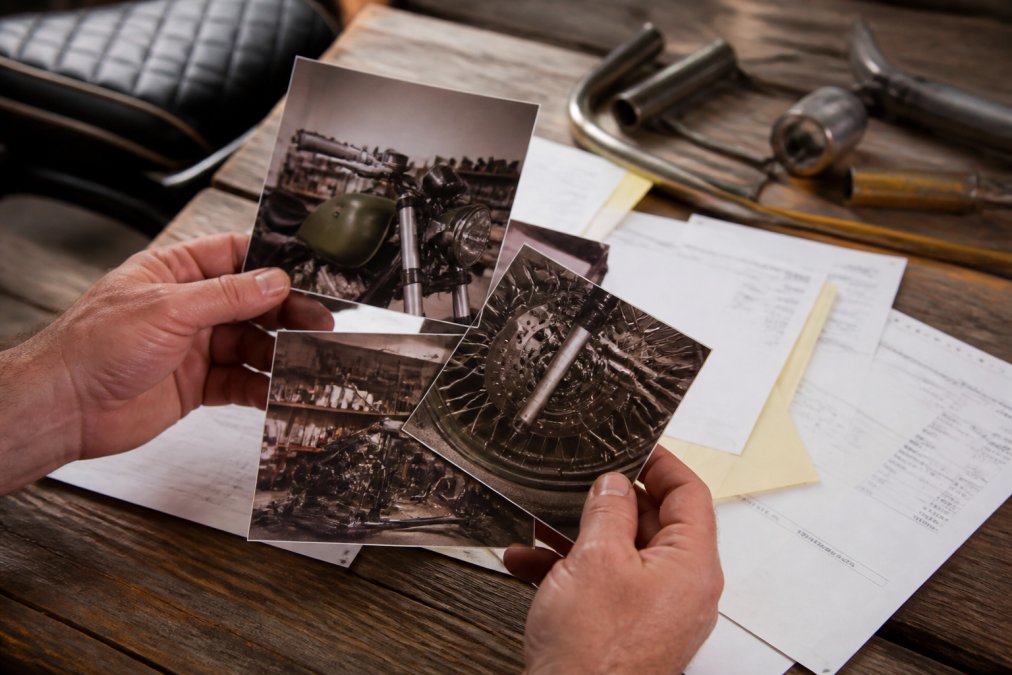 Close-up of hands holding photographs and receipts of a custom motorcycle build, with parts documentation and fabrication invoices spread across a wooden table in natural daylight