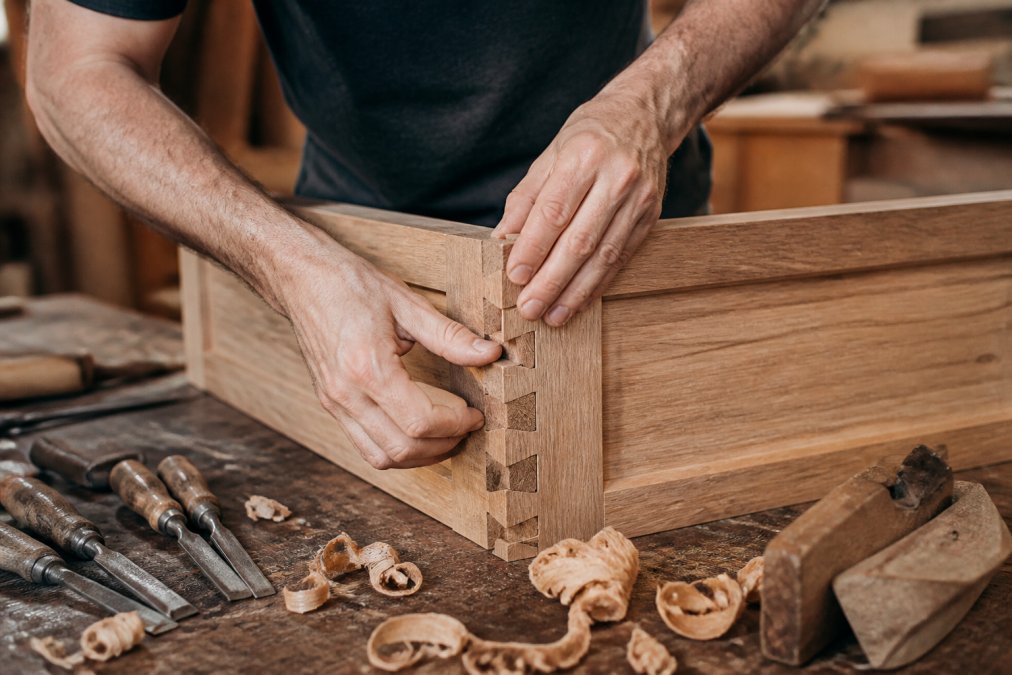 Close-up of a craftsman's hands carefully fitting a dovetail joint on a solid oak cabinet door in a traditional joinery workshop, wood shavings and chisels arranged on the workbench beside the piece
