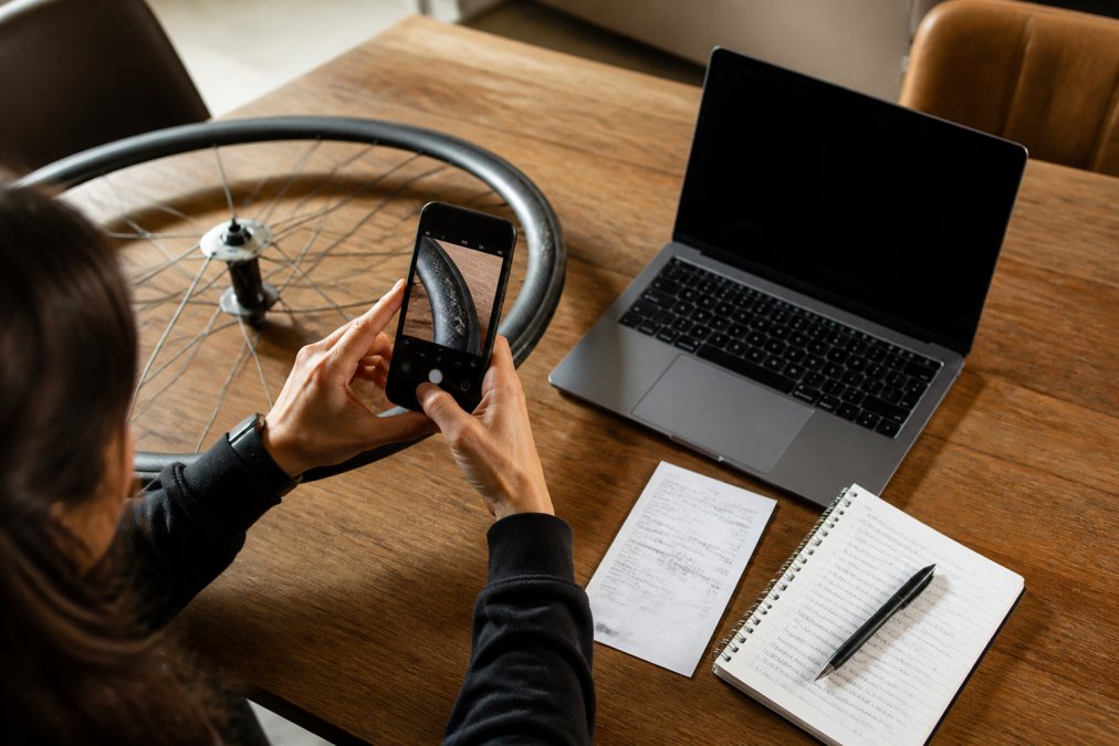 Person seated at a timber desk photographing a damaged carbon wheelset with a smartphone, laptop open to a warranty claim form, printed receipt and handwritten notes visible in warm indoor light