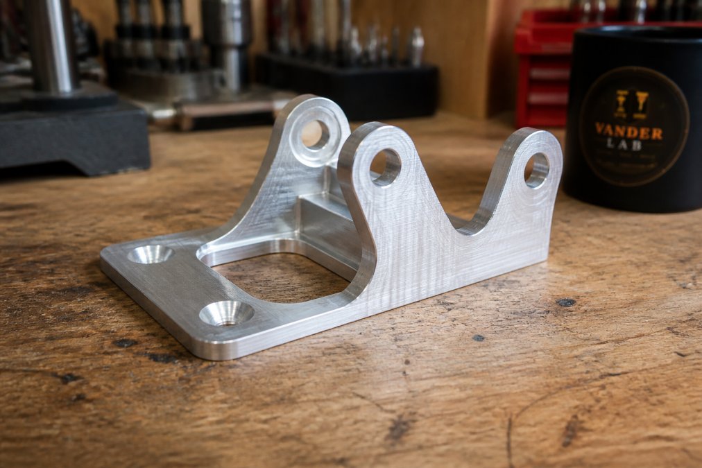 Close-up of a finished CNC-machined aluminum bracket sitting on a workbench in a home fabrication lab, showing crisp edges and a smooth machined surface under bright task lighting