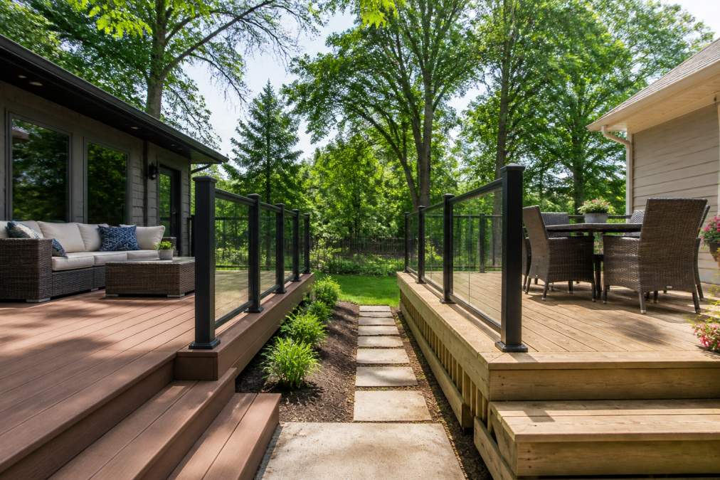 Side-by-side view of two residential decks in a sunny backyard with mature trees, one built with warm-toned composite boards and the other with natural wood grain, both featuring modern glass and metal railing systems