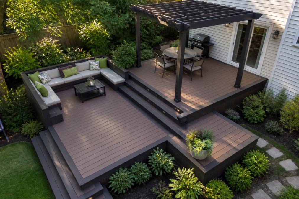 Overhead view of a custom multi-level deck in a small backyard showing elevated composite decking platforms, built-in wraparound seating, and a pergola structure creating distinct outdoor zones with natural afternoon lighting