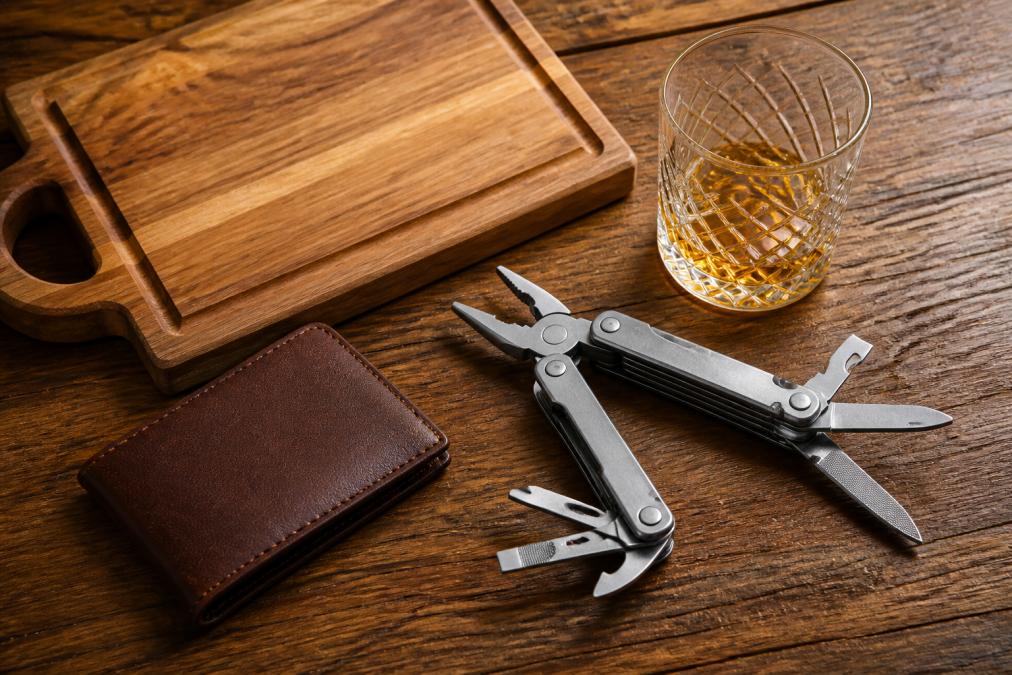 Flat lay of assorted personalized gifts for dad, engraved whiskey glass, wooden cutting board, multi-tool, and leather wallet arranged on a wooden surface with warm lighting