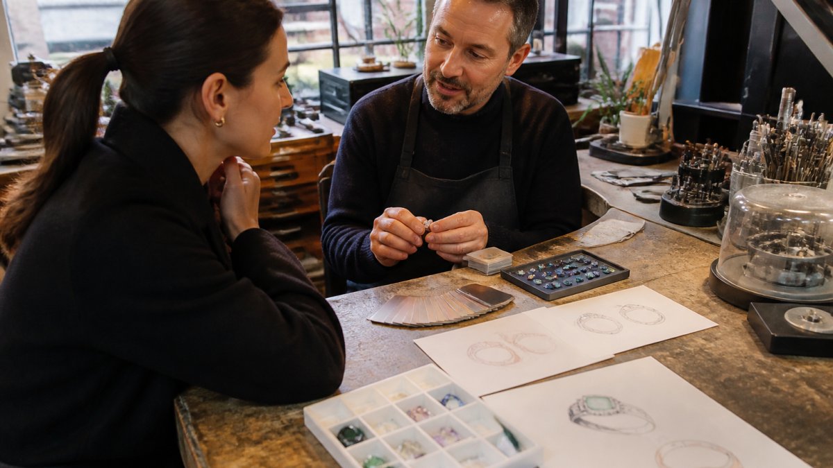 Professional jewelry designer and client in conversation at workbench surrounded by gemstone samples, metal swatches, and inspiration sketches, examining design references together under warm studio lighting