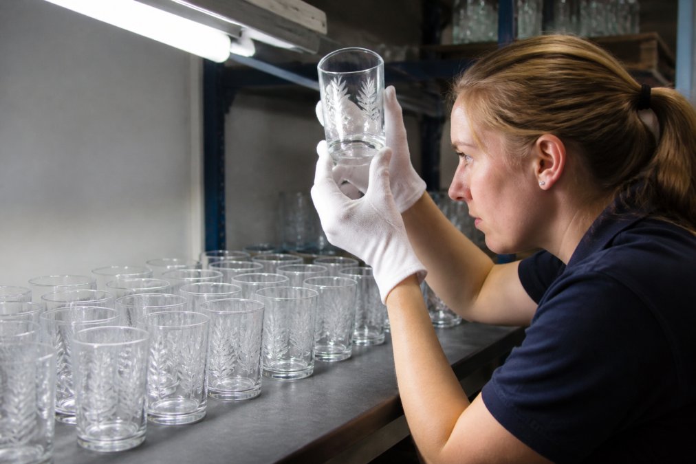 Quality control technician inspecting custom etched glassware under bright lighting, examining surface finish and engraving clarity on multiple samples