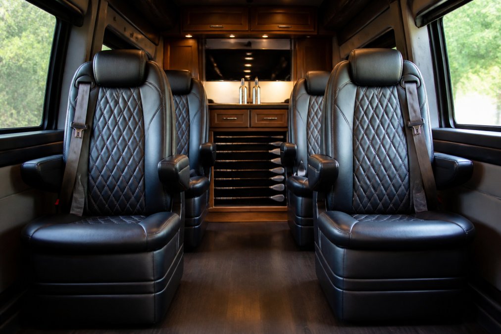 Interior of a luxury conversion van showing four plush leather captain's chairs, custom wood cabinetry, and a secure horizontal golf club storage rack mounted near the floor with soft ambient lighting