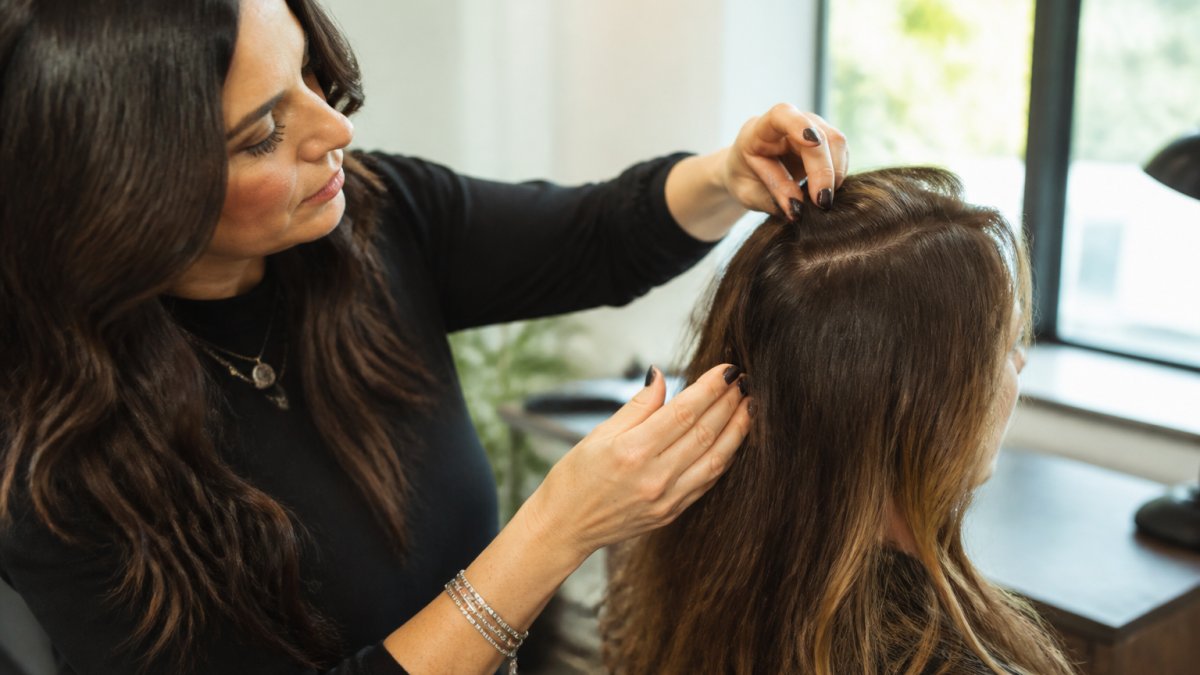 Professional stylist examining client's natural hair texture and density at crown and part area during consultation, natural lighting in private studio