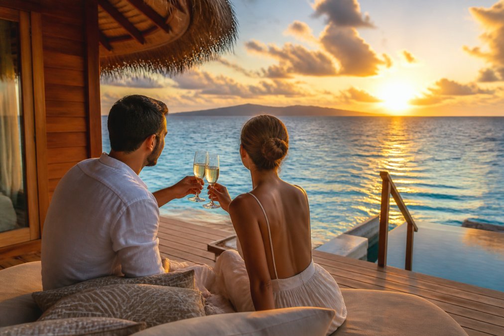 A couple toasting champagne on a private overwater bungalow deck at sunset, with a tropical ocean view stretching behind them, warm golden light reflecting off the water
