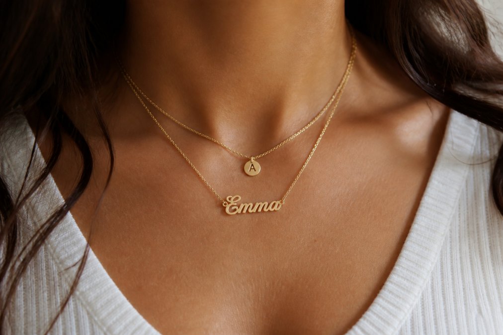 Close-up of a woman's neck wearing a layered necklace stack featuring a dainty initial pendant and a nameplate necklace, soft natural lighting against a neutral top