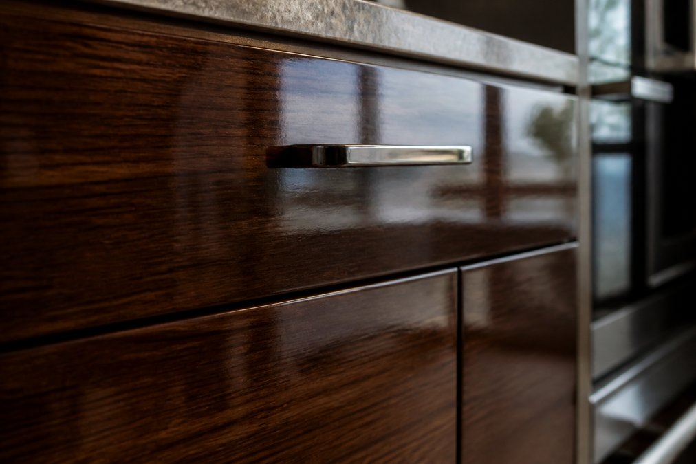 Close-up of Italian kitchen cabinet door showing glossy lacquer finish with rich colour depth, premium metal hardware catch reflecting soft natural light, and wood grain detail visible through translucent finish