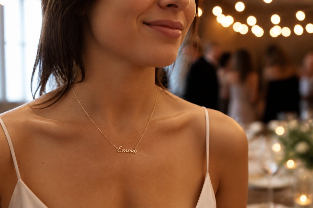 Close-up of a woman's neck wearing a delicate gold cursive name necklace, soft window light on her collarbone, warm blurred celebration background with string lights
