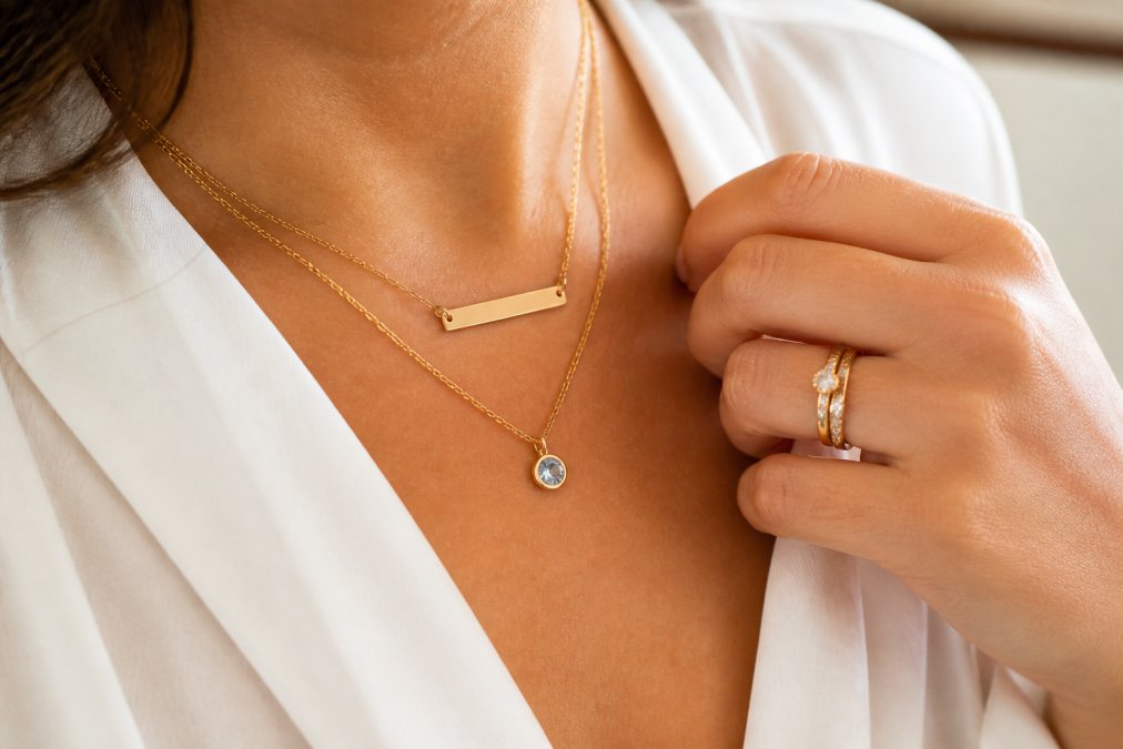 Close-up of a woman's hand and neckline wearing a delicate gold nameplate necklace and a birthstone pendant, resting against a soft neutral fabric background with warm lighting