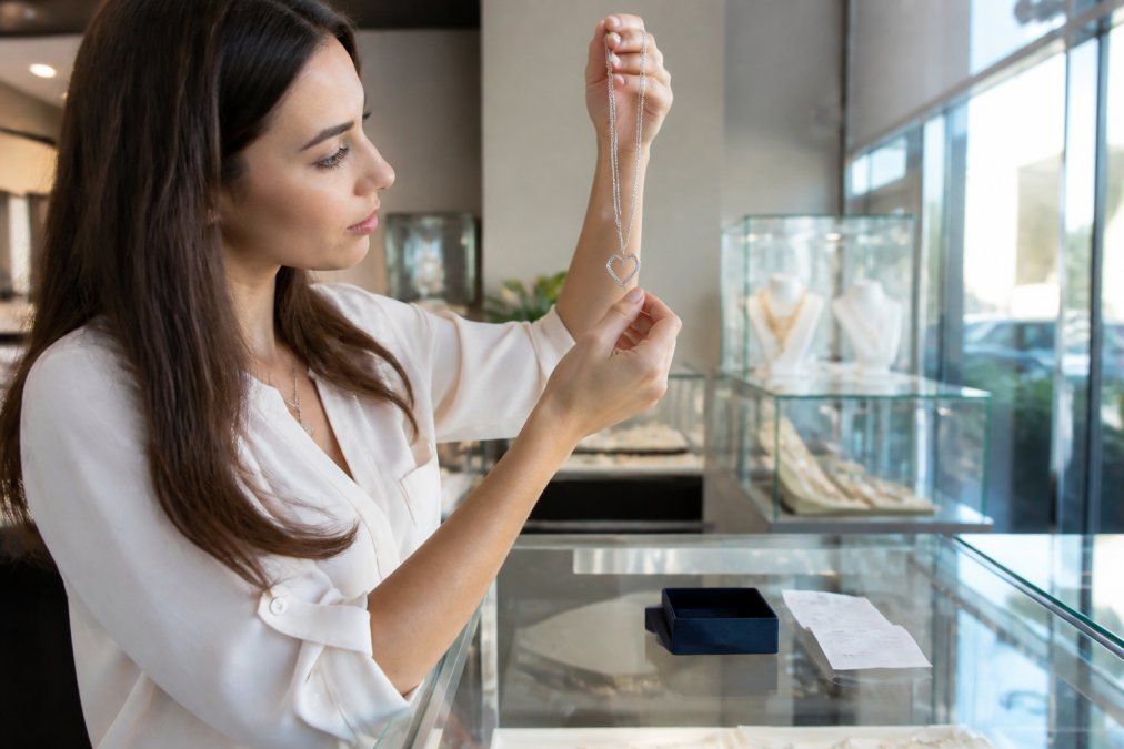 A customer carefully inspecting a personalized necklace in a bright jewelry store, holding the piece up to the light with a receipt and small jewelry box on the counter nearby