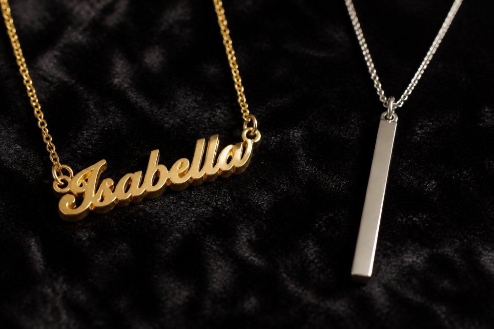 Close-up of two necklaces side by side on a dark velvet surface, one handcrafted name necklace with visible artisan detail and heavier metal weight, one generic uniform pendant with a thin, machine-stamped finish, under warm studio lighting