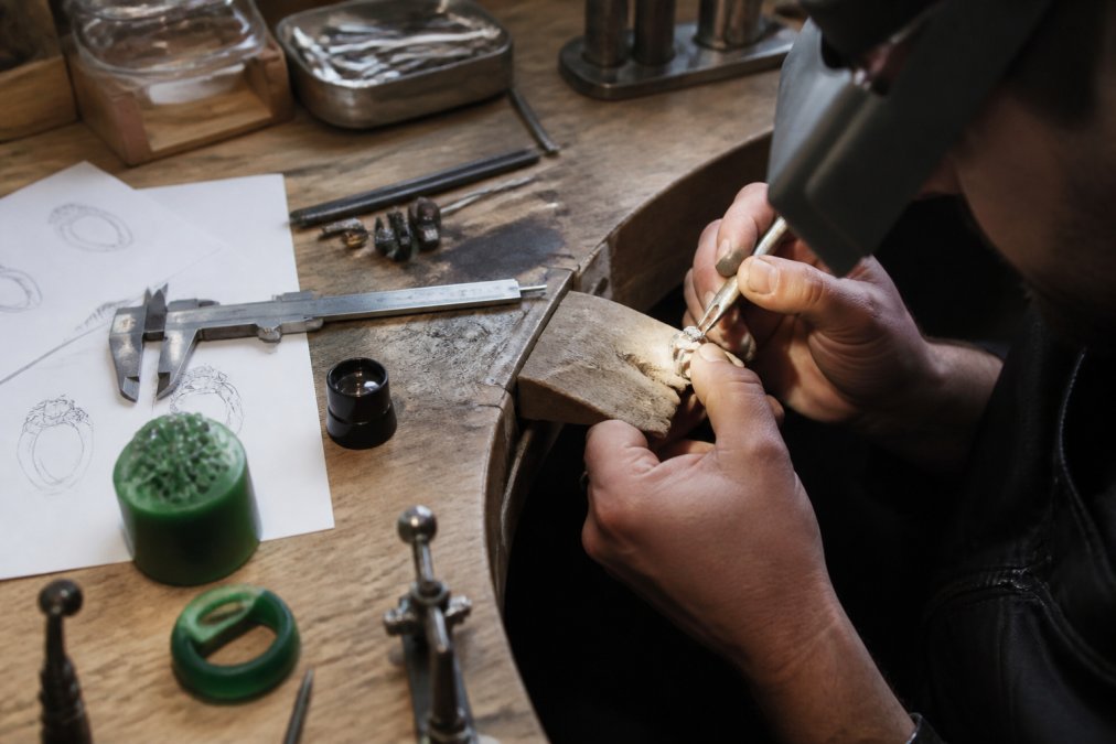 A bench jeweler's hands working at a lit workbench, setting a stone into a ring with fine tools, wax carving and CAD sketches visible on the bench beside a loupe and calipers